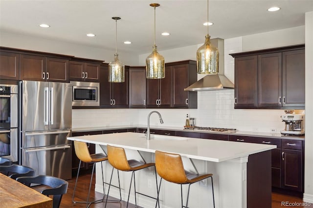 kitchen featuring a kitchen island with sink, a breakfast bar, wall chimney range hood, a sink, and stainless steel appliances