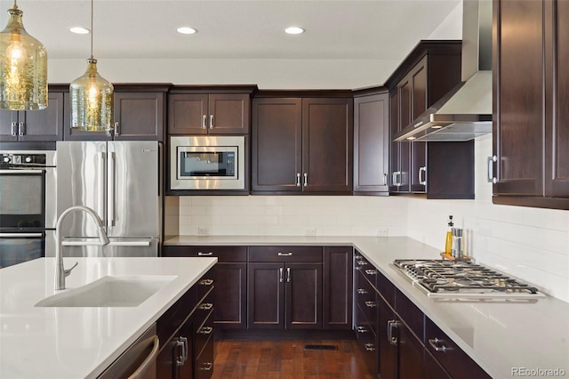 kitchen with wall chimney range hood, dark brown cabinetry, appliances with stainless steel finishes, a sink, and light countertops