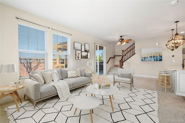 living room with ceiling fan with notable chandelier, stairway, light colored carpet, and baseboards