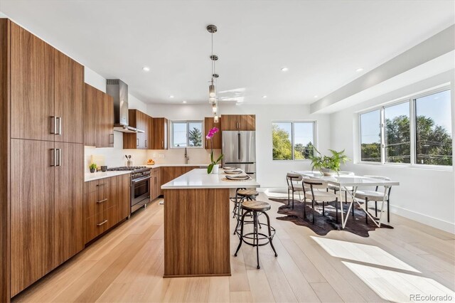 kitchen featuring wall chimney exhaust hood, light wood-type flooring, decorative light fixtures, appliances with stainless steel finishes, and a center island