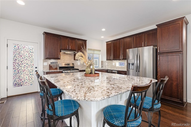 kitchen featuring a center island, light stone countertops, under cabinet range hood, stainless steel appliances, and dark wood-type flooring