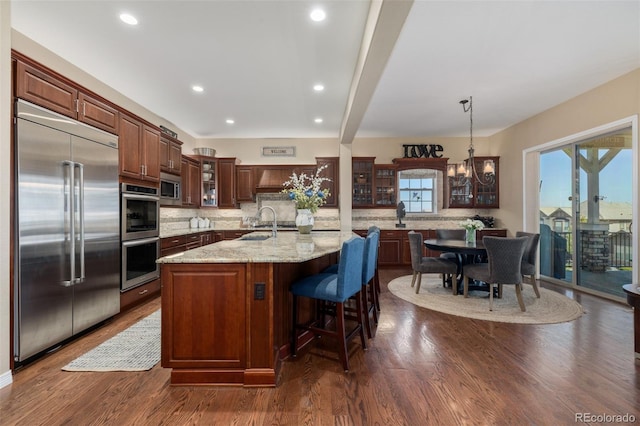 kitchen featuring a kitchen island with sink, built in appliances, backsplash, and glass insert cabinets