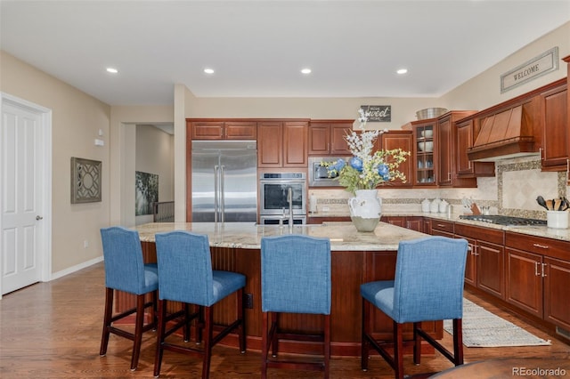 kitchen featuring light stone countertops, built in appliances, an island with sink, backsplash, and glass insert cabinets