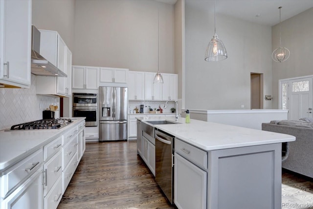 kitchen featuring appliances with stainless steel finishes, a high ceiling, white cabinets, wall chimney exhaust hood, and a sink