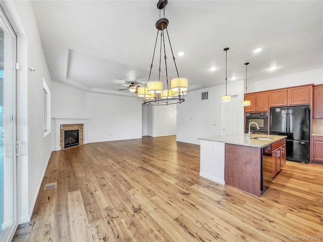 kitchen with decorative light fixtures, a tile fireplace, black appliances, a kitchen island with sink, and light stone counters