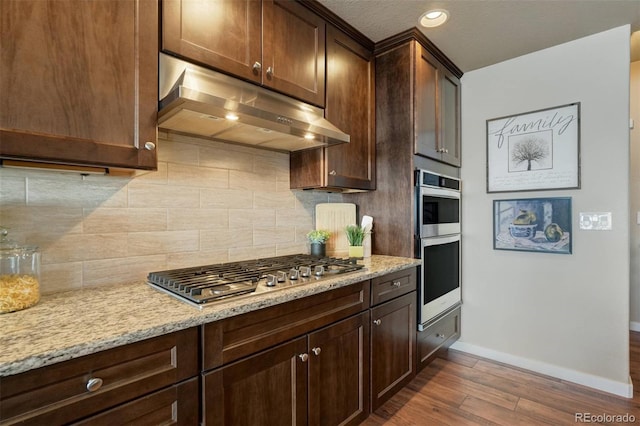 kitchen featuring light stone counters, under cabinet range hood, dark wood-type flooring, appliances with stainless steel finishes, and decorative backsplash