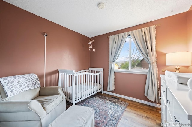 bedroom featuring a crib, wood finished floors, visible vents, baseboards, and a textured ceiling