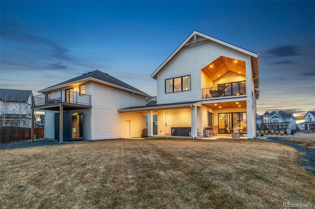 back house at dusk featuring a lawn, a balcony, a patio, and a hot tub