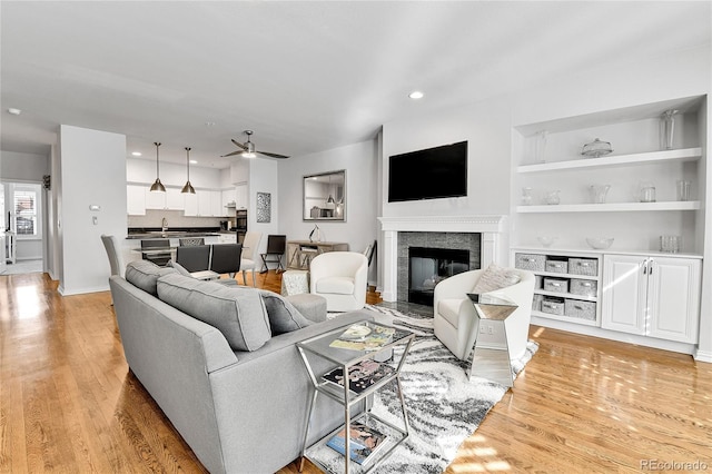 living room with light wood finished floors, ceiling fan, a fireplace with flush hearth, built in features, and recessed lighting