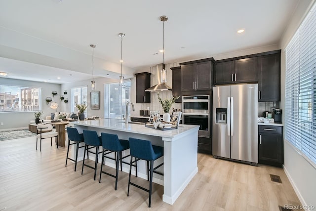 kitchen with appliances with stainless steel finishes, tasteful backsplash, wall chimney range hood, pendant lighting, and an island with sink