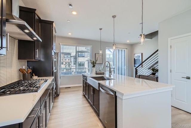 kitchen with wall chimney range hood, an island with sink, appliances with stainless steel finishes, and pendant lighting