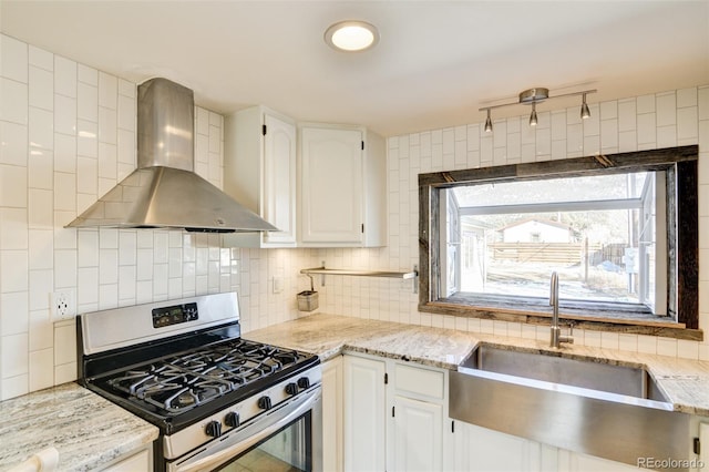 kitchen with decorative backsplash, white cabinets, stainless steel gas range, a sink, and wall chimney range hood