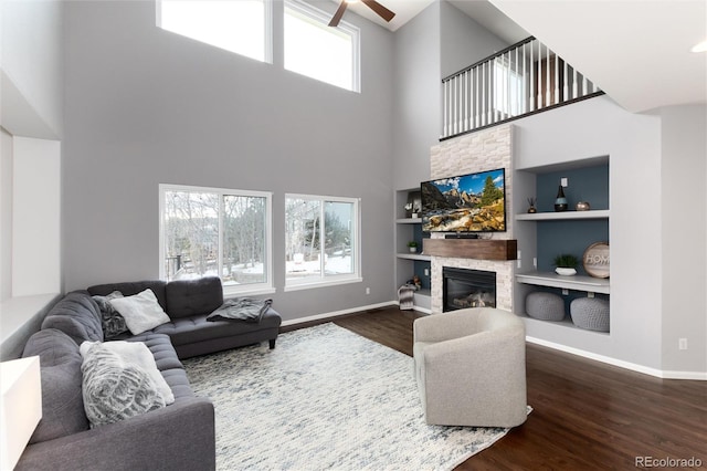 living room with a towering ceiling, a fireplace, built in shelves, dark wood-type flooring, and ceiling fan