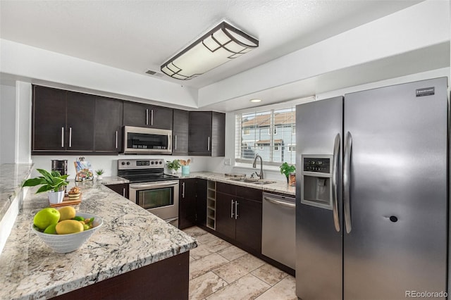 kitchen with dark brown cabinetry, light stone countertops, sink, kitchen peninsula, and appliances with stainless steel finishes