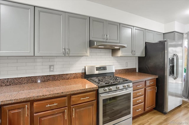 kitchen with brown cabinetry, light wood-style flooring, appliances with stainless steel finishes, under cabinet range hood, and backsplash