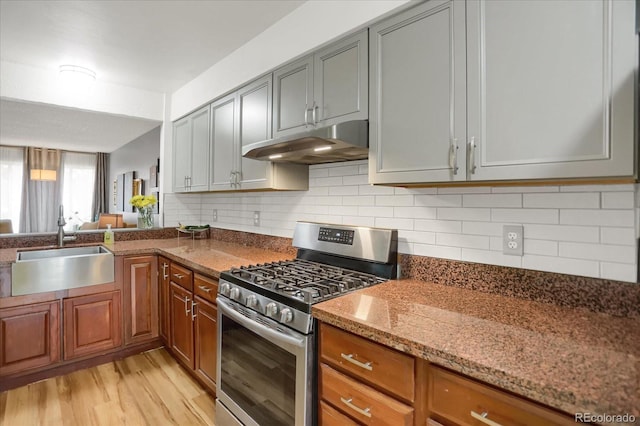 kitchen with stainless steel gas range oven, under cabinet range hood, a sink, dark stone counters, and light wood finished floors