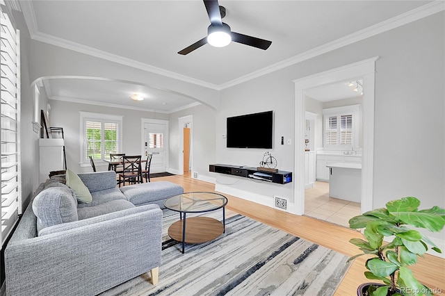 living area with ornamental molding, baseboards, ceiling fan, arched walkways, and light wood finished floors