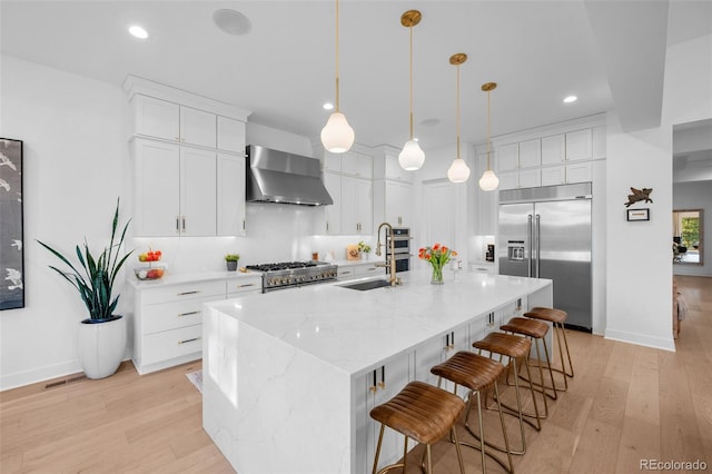 kitchen featuring a sink, wall chimney range hood, built in fridge, light wood finished floors, and stove