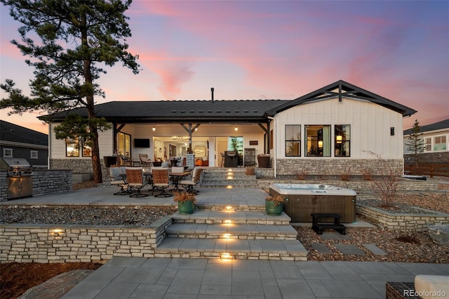 back of house at dusk featuring area for grilling, stone siding, a patio area, board and batten siding, and a hot tub