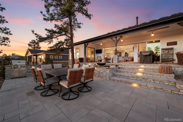 patio terrace at dusk featuring outdoor dining space, an outdoor kitchen, a fire pit, and grilling area