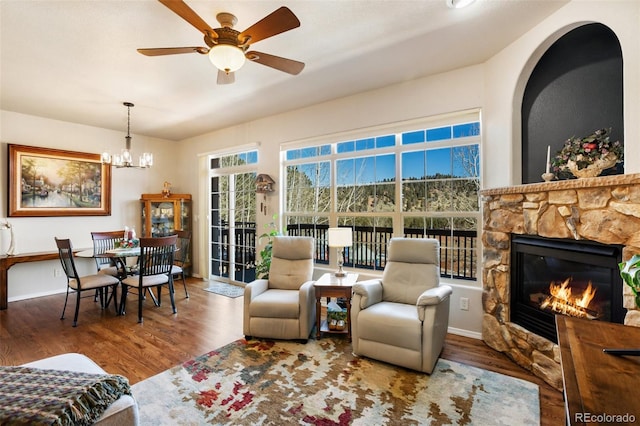 living room with baseboards, a stone fireplace, wood finished floors, and ceiling fan with notable chandelier