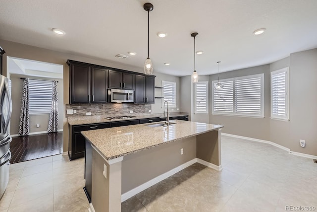 kitchen featuring an island with sink, sink, hanging light fixtures, stainless steel appliances, and light stone countertops