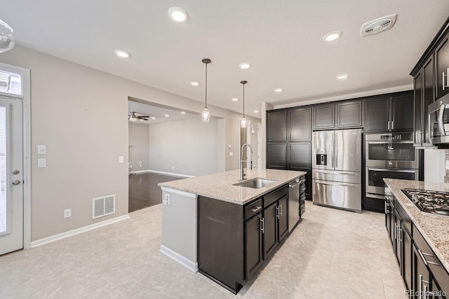 kitchen featuring sink, a kitchen island with sink, hanging light fixtures, stainless steel appliances, and light stone counters