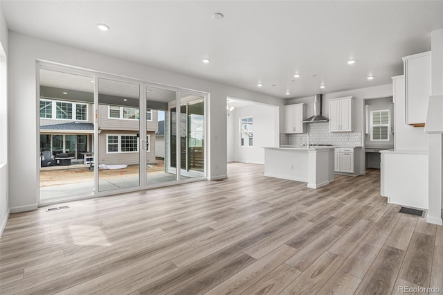 kitchen with a kitchen island with sink, light wood-type flooring, white cabinetry, tasteful backsplash, and wall chimney range hood