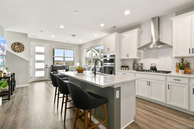 kitchen with white cabinets, appliances with stainless steel finishes, wood-type flooring, wall chimney range hood, and a center island with sink