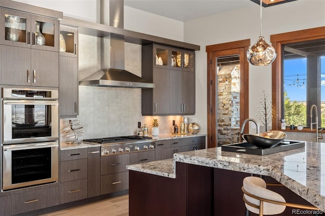 kitchen with light stone counters, hanging light fixtures, wall chimney range hood, light hardwood / wood-style flooring, and stainless steel appliances