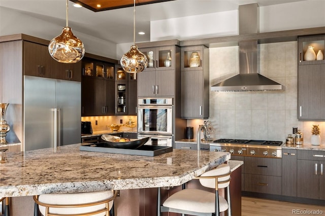 kitchen featuring a breakfast bar, light hardwood / wood-style floors, wall chimney exhaust hood, stainless steel appliances, and dark brown cabinetry