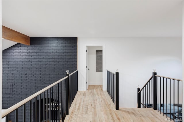 hallway featuring light wood-type flooring, beam ceiling, brick wall, and an upstairs landing