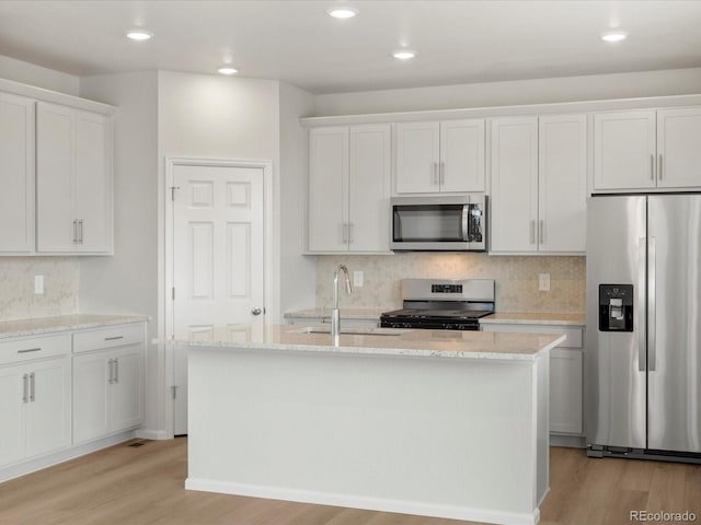 kitchen featuring appliances with stainless steel finishes, a sink, light wood finished floors, a center island with sink, and decorative backsplash