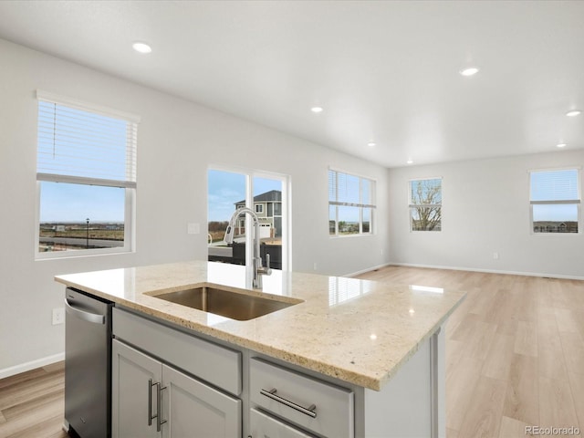 kitchen with a sink, stainless steel dishwasher, a center island with sink, light wood-style flooring, and light stone countertops