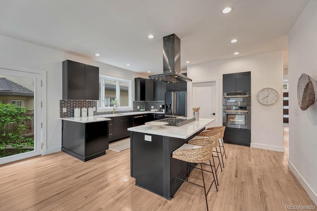 kitchen featuring island exhaust hood, stainless steel refrigerator with ice dispenser, dark cabinetry, decorative backsplash, and a kitchen breakfast bar