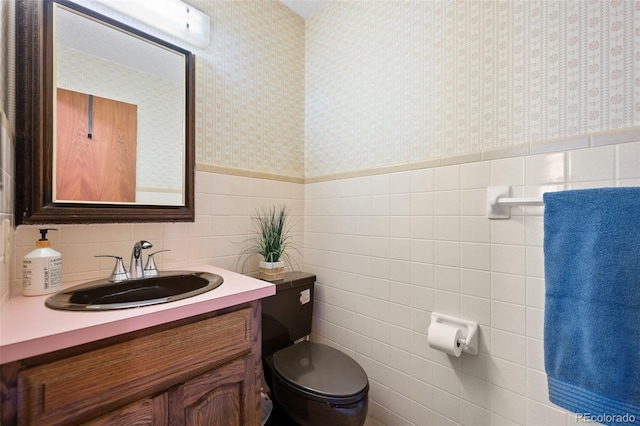 half bath with tile walls, toilet, wainscoting, vanity, and wallpapered walls