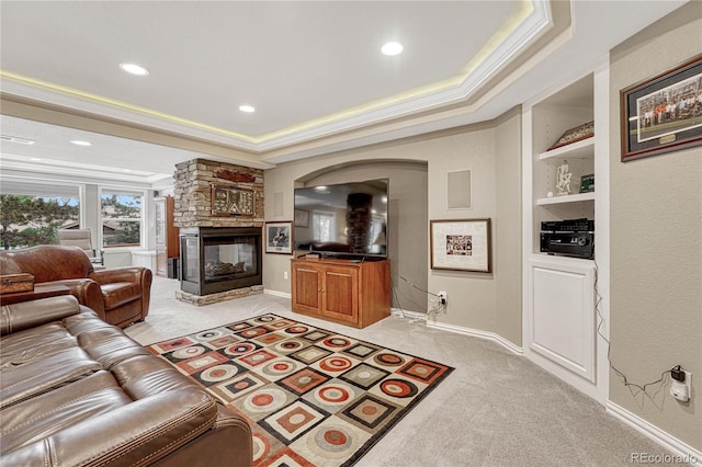 carpeted living room with a stone fireplace, built in shelves, a raised ceiling, and crown molding