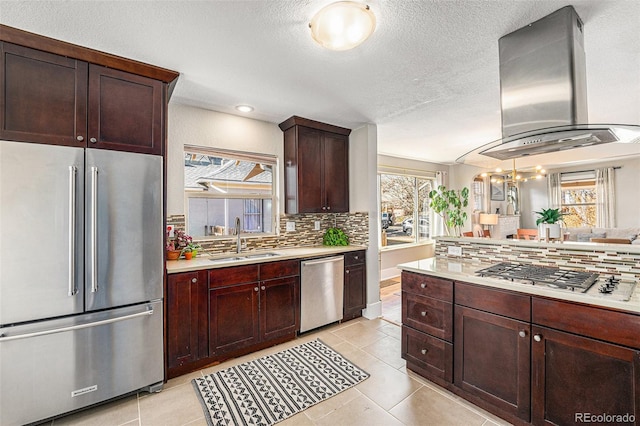 kitchen featuring tasteful backsplash, island range hood, appliances with stainless steel finishes, light countertops, and a sink