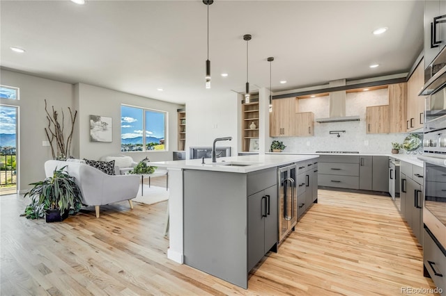 kitchen with light brown cabinetry, a center island with sink, wall chimney exhaust hood, and light wood-type flooring