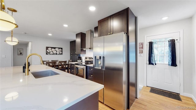 kitchen featuring dark brown cabinets, light wood-style flooring, appliances with stainless steel finishes, wall chimney exhaust hood, and a sink