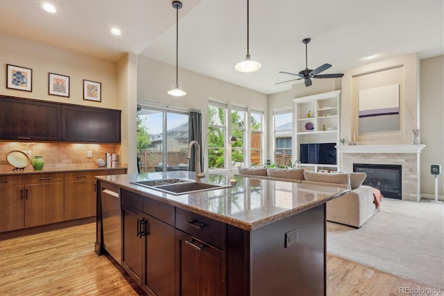 kitchen with a sink, stainless steel dishwasher, a ceiling fan, a glass covered fireplace, and a center island with sink