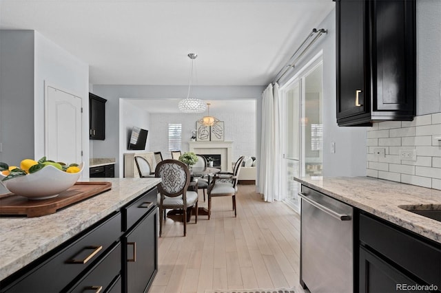 kitchen with dark cabinets, stainless steel dishwasher, backsplash, light stone countertops, and decorative light fixtures