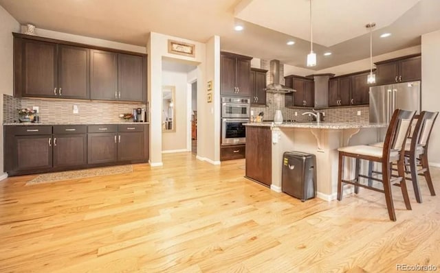 6765 Windbrook Ct, Colorado Springs, CO 80927 - kitchen featuring wall chimney range hood, stainless steel appliances, tasteful backsplash, light wood-style floors, and a breakfast bar area