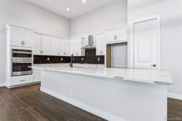 kitchen with dark wood-type flooring, white cabinets, a center island with sink, wall chimney exhaust hood, and light stone countertops