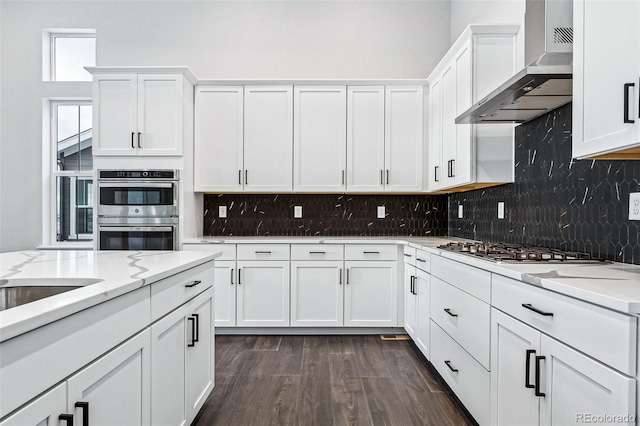 kitchen with dark wood-type flooring, white cabinets, wall chimney range hood, light stone countertops, and appliances with stainless steel finishes