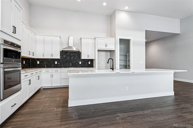 kitchen with stainless steel double oven, a kitchen island with sink, dark wood-type flooring, wall chimney range hood, and white cabinetry