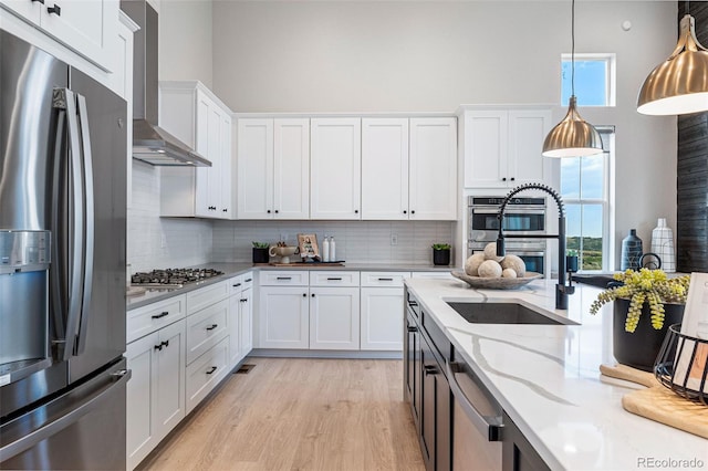 kitchen with light stone countertops, stainless steel appliances, wall chimney range hood, pendant lighting, and white cabinetry