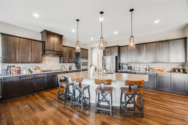 kitchen featuring a center island with sink, stainless steel appliances, dark wood-type flooring, dark brown cabinets, and a kitchen bar