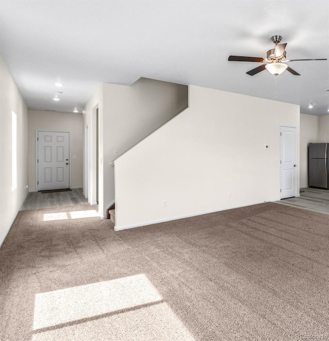 unfurnished living room featuring stairs, light colored carpet, baseboards, and ceiling fan