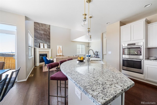 kitchen with light stone counters, an island with sink, stainless steel double oven, dark wood-type flooring, and a sink